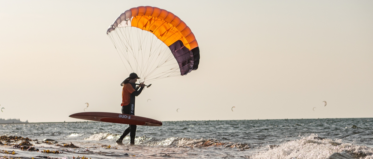 Parawing_gegenlicht Foiler mit Foilboard auf dem Arm geht zum Wasser und hält dabei den Parawing in der Hand